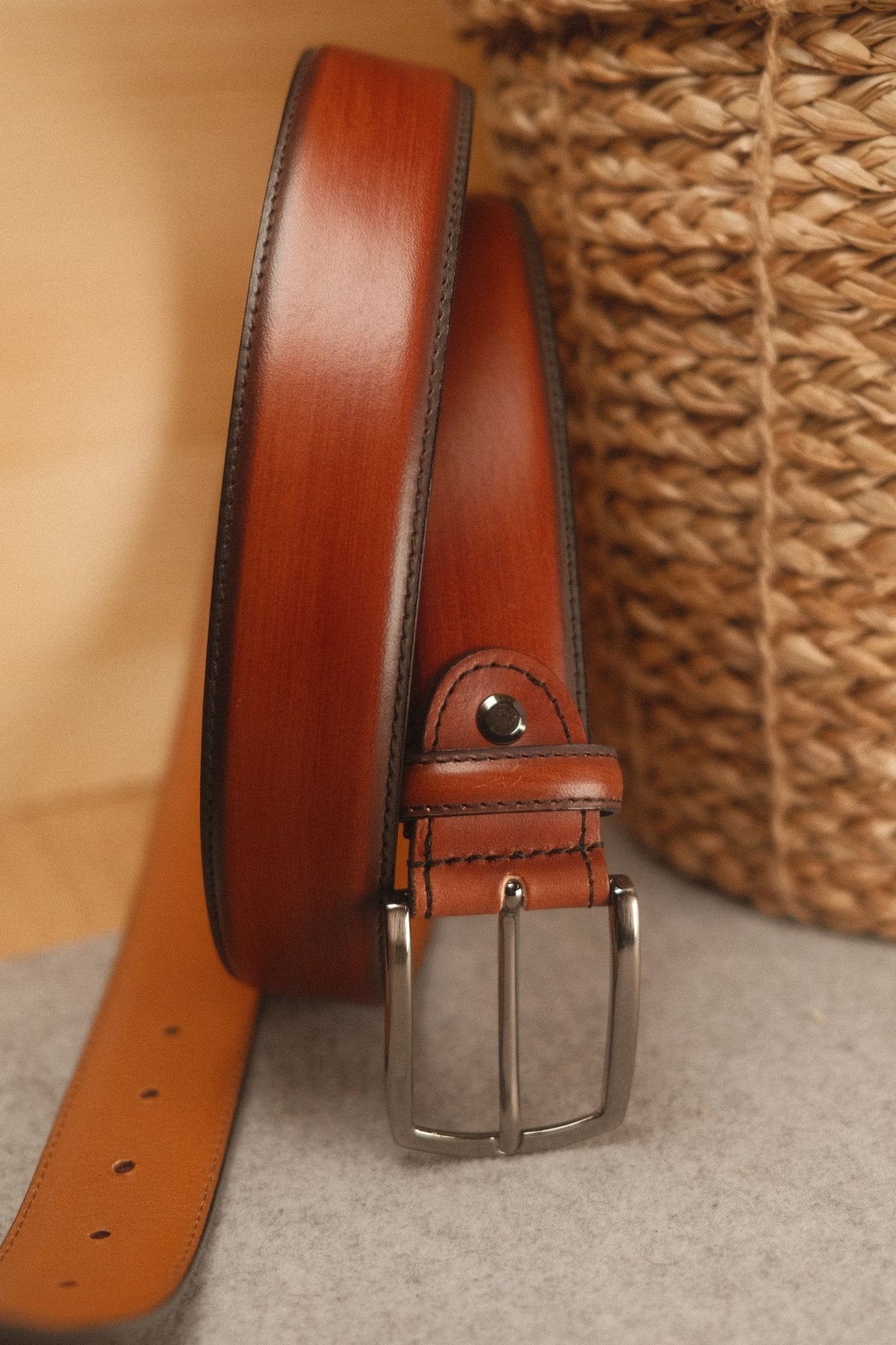 Ceinture en cuir patiné cognac roulée devant un panier tressé : la boucle, son ardillon, la vis et les trous de la pointe.