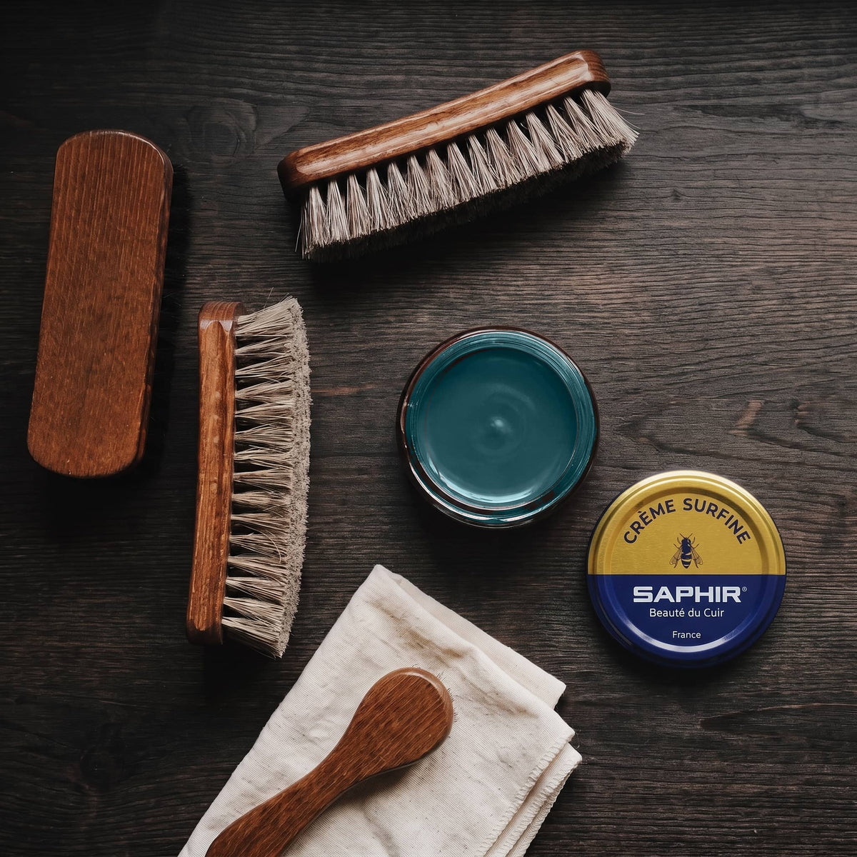 Crème Surfine Saphir Vert foncé en pot ouvert, avec des brosses en crin et une chamoisine, vue de dessus